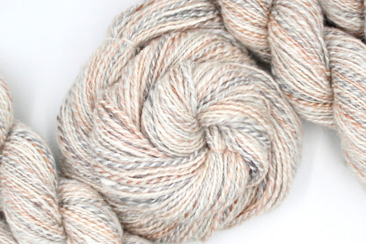 A one of a kind, hand dyed variegated skein of multicolored Cream, Dusky Pink, and Grey self-striping wool Yarn coiled attractively in the center of the frame.