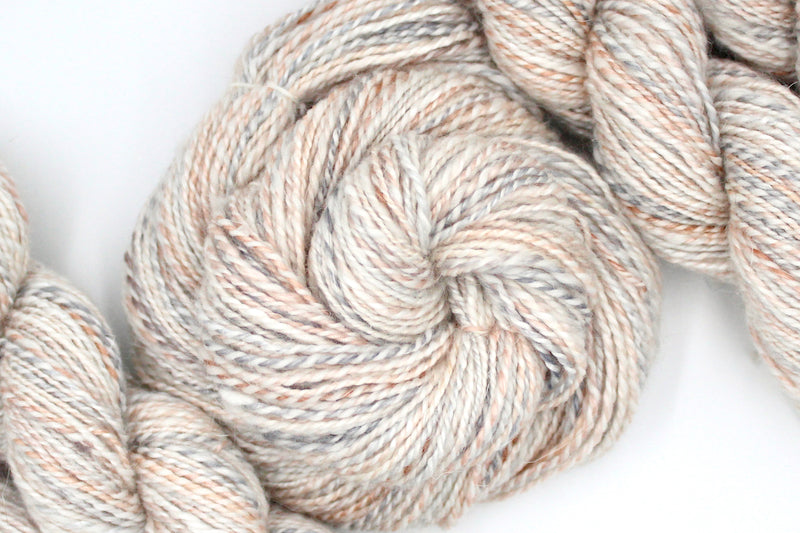A one of a kind, hand dyed variegated skein of multicolored Cream, Dusky Pink, and Grey self-striping wool Yarn coiled attractively in the center of the frame. 