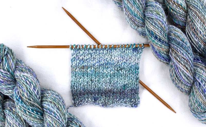 A sample swatch knitted from a one of a kind, hand dyed variegated skein of multicolored Navy, Teal, Royal Blue, Lavender, and Tan self-striping wool Yarn. 