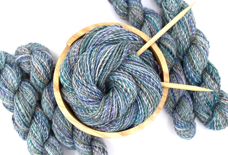 A one of a kind, hand dyed variegated skein of multicolored Navy, Teal, Royal Blue, Lavender, and Tan self-striping wool Yarn, in a yarn bowl with knitting needles, ready to be made into something beautiful! 