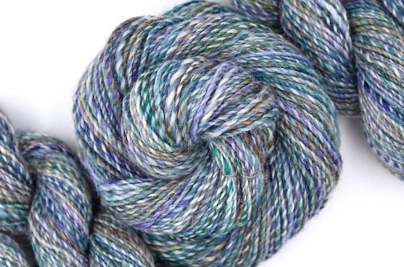 A one of a kind, hand dyed variegated skein of multicolored Navy, Teal, Royal Blue, Lavender, and Tan self-striping wool Yarn coiled attractively in the center of the frame. 