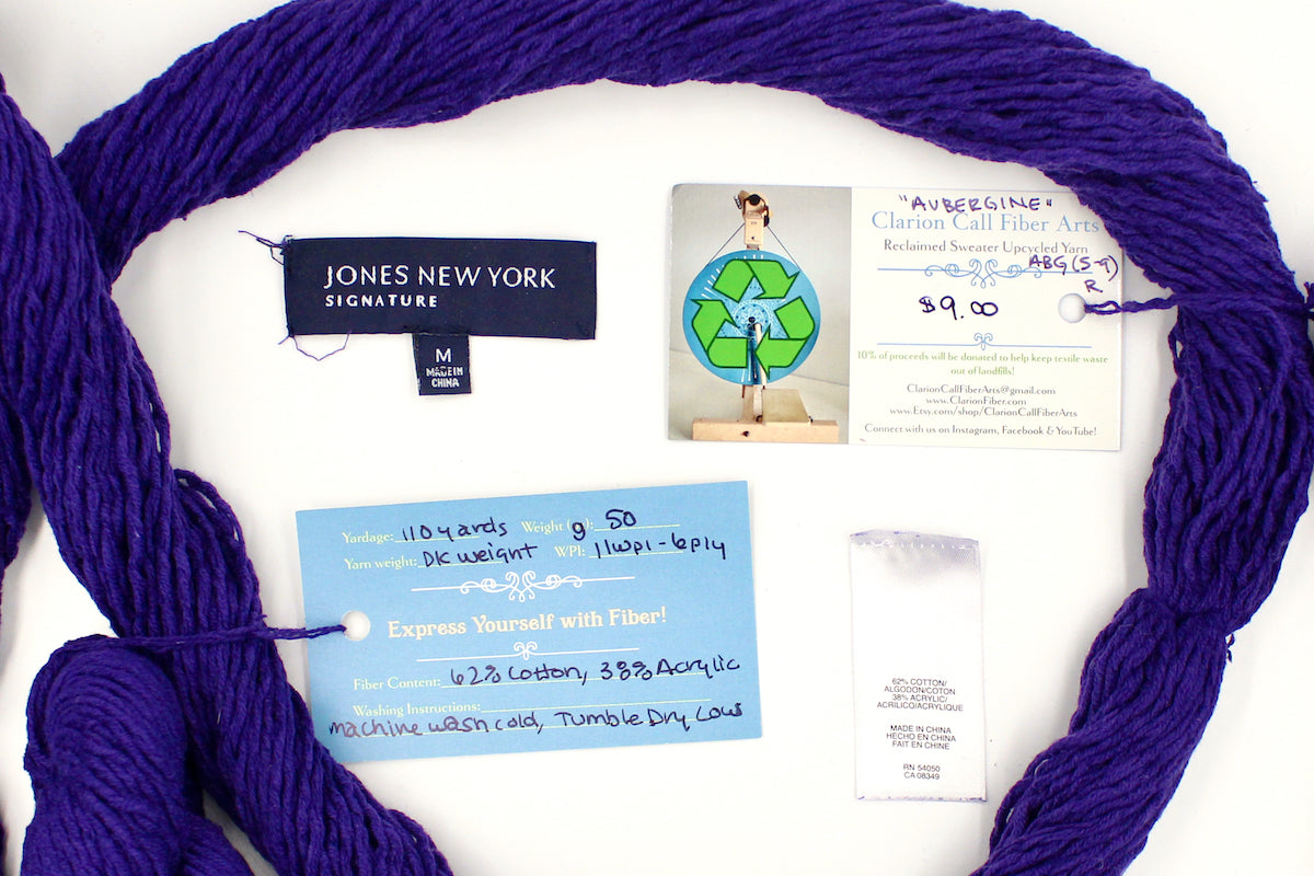 A skein of Vegan, Purple, 62% Cotton, 38% Acrylic, DK Weight yarn encircling the sweater labels it was recycled from, as well as the hand written yarn tags detailing the specs of this recycled yarn to help you make your next project! 