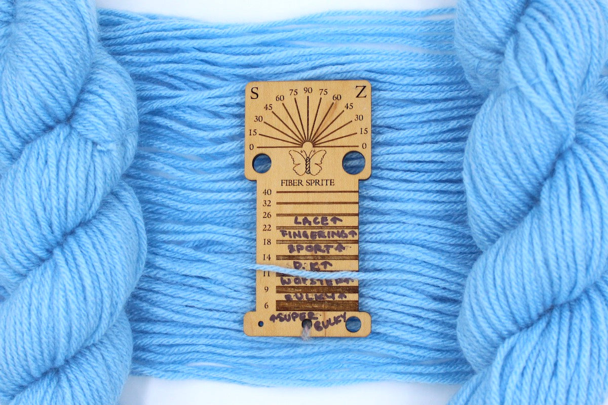 A skein of sport weight baby blue Yarn recycled by hand from unwanted sweaters draped over a Yarn weight tool