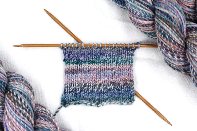 A sample swatch knitted from a one of a kind, hand dyed variegated skein of multicolored Blue, Teal, Pink, Brown and Tan self-striping wool Yarn. 