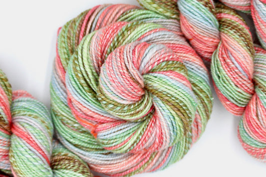 A one of a kind, hand dyed gradient skein of multicolored Pink, Blue, Green, Tan, and Brown self-striping wool Yarn coiled attractively in the center of the frame. 