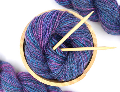 A one of a kind, hand dyed Variegated skein of multicolored Hot Pink, Blue, and Purple self-striping wool Yarn, in a yarn bowl with knitting needles, ready to be made into something beautiful! 