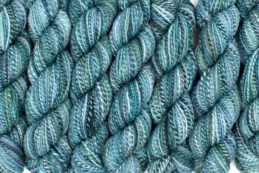 Six one of a kind, DK Weight, hand dyed variegated skeins of multicolored Blue, Teal, and Green self-striping 100% Merino wool Yarn lined up side by side. 