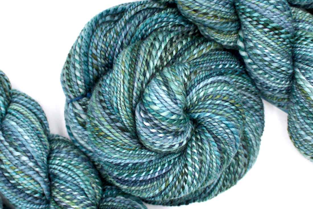 A one of a kind, DK Weight, hand dyed variegated skein of multicolored Blue, Teal, and Green self-striping 100% Merino wool Yarn coiled attractively in the center of the frame. 