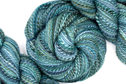 A one of a kind, DK Weight, hand dyed variegated skein of multicolored Blue, Teal, and Green self-striping 100% Merino wool Yarn coiled attractively in the center of the frame. 