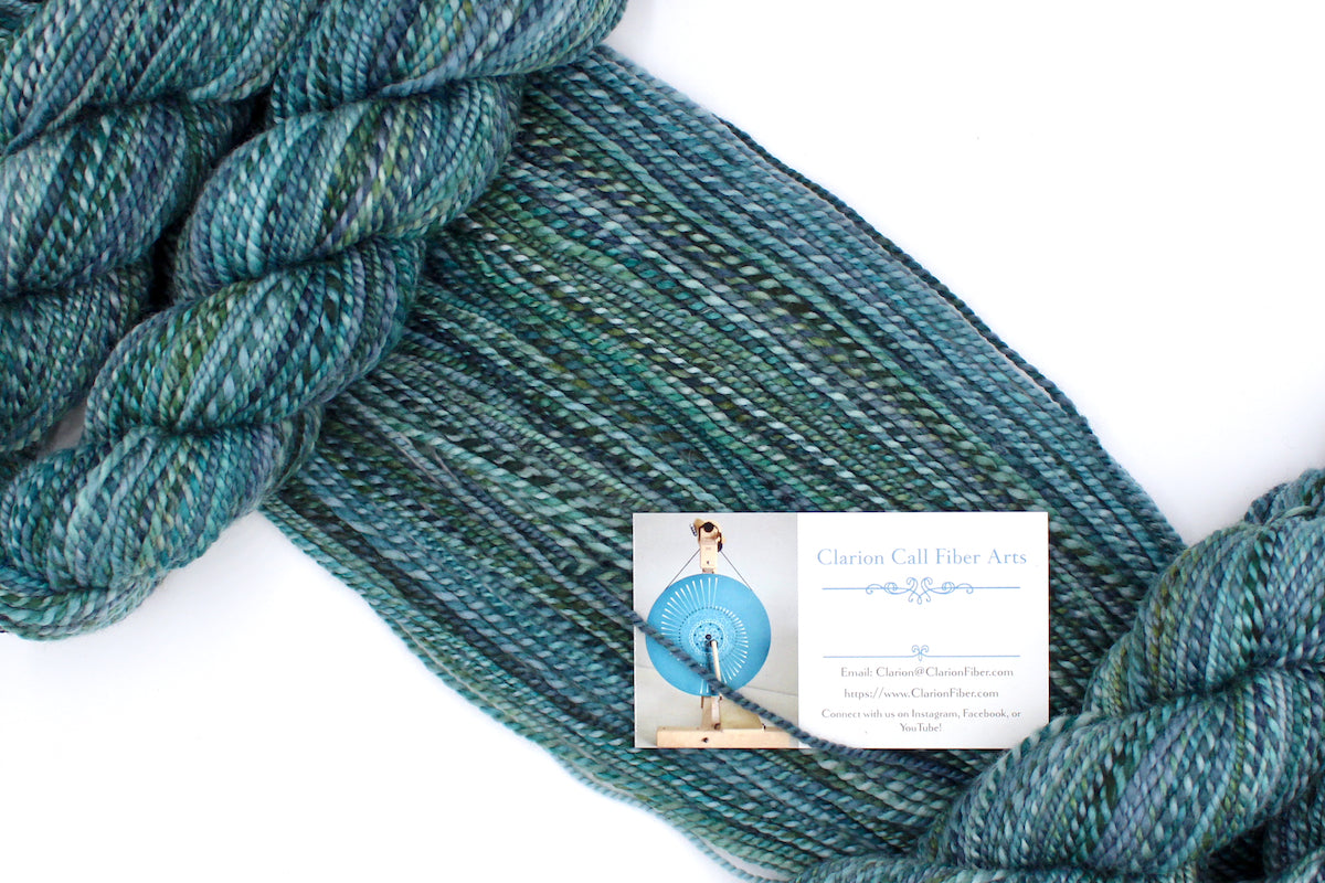 A one of a kind, DK Weight, hand dyed variegated skein of multicolored Blue, Teal, and Green self-striping 100% Merino wool Yarn draped diagonally across the frame, so you can really see the color play. 