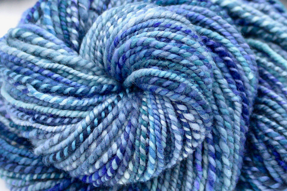 A close up view of a one of a kind, worsted Weight, hand dyed variegated skein of multicolored Blue and Purple self-striping Hand Spun Yarn, made of 100% Hand Dyed Merino Wool. 