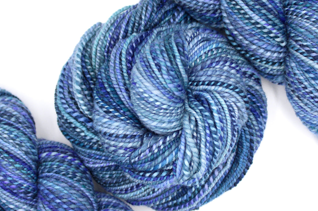 A one of a kind, worsted Weight, hand dyed variegated skein of multicolored Blue and Purple self-striping Hand Spun Yarn, made of 100% Hand Dyed Merino Wool and coiled attractively in the center of the frame. 