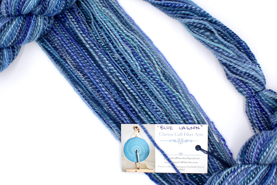 A one of a kind, worsted Weight, hand dyed variegated skein of multicolored Blue and Purple self-striping Hand Spun Yarn made of 100% Hand Dyed Merino Wool, draped diagonally across the frame, so you can really see the color play. 