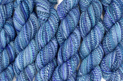 Six one of a kind, worsted Weight, hand dyed variegated skeins of multicolored Blue and Purple self-striping Hand Spun Yarn, made of 100% Hand Dyed Merino Wool lined up side by side. 