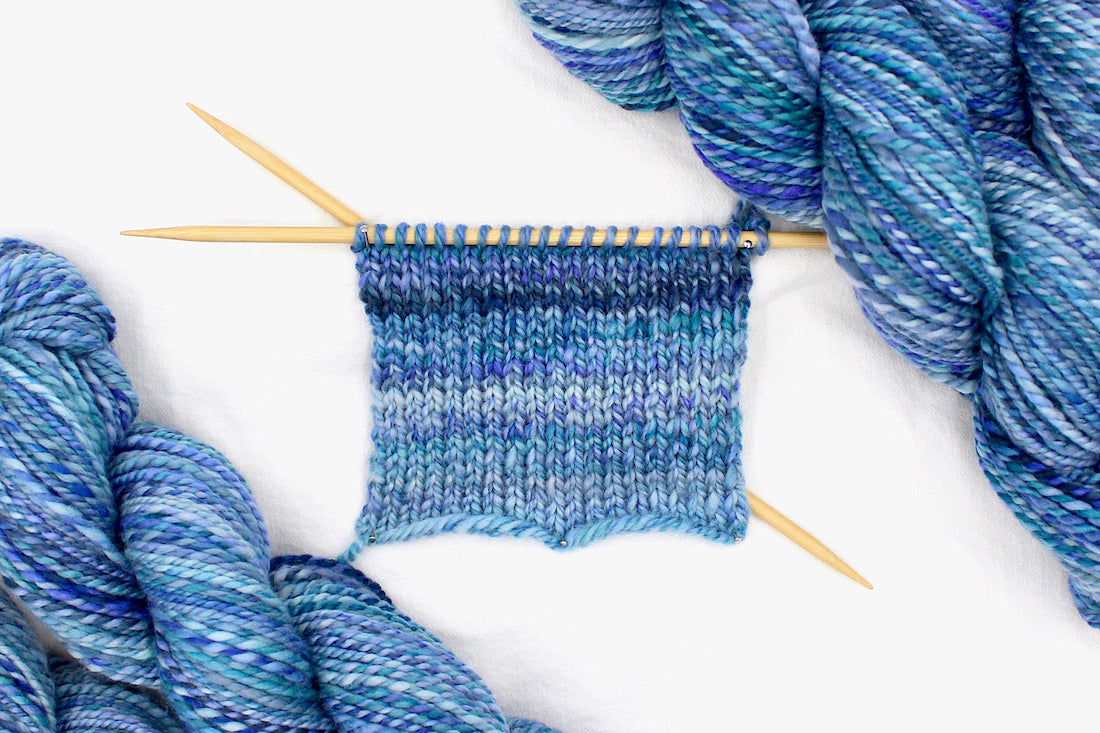 A sample swatch knitted on size 5 knitting needles from a one of a kind, worsted Weight, hand dyed variegated skein of multicolored Blue and Purple self-striping Hand Spun Yarn, made of 100% Hand Dyed Merino Wool. 