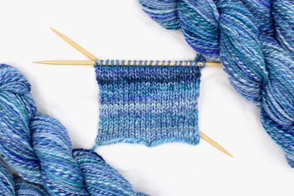 A sample swatch knitted on size 5 knitting needles from a one of a kind, worsted Weight, hand dyed variegated skein of multicolored Blue and Purple self-striping Hand Spun Yarn, made of 100% Hand Dyed Merino Wool. 
