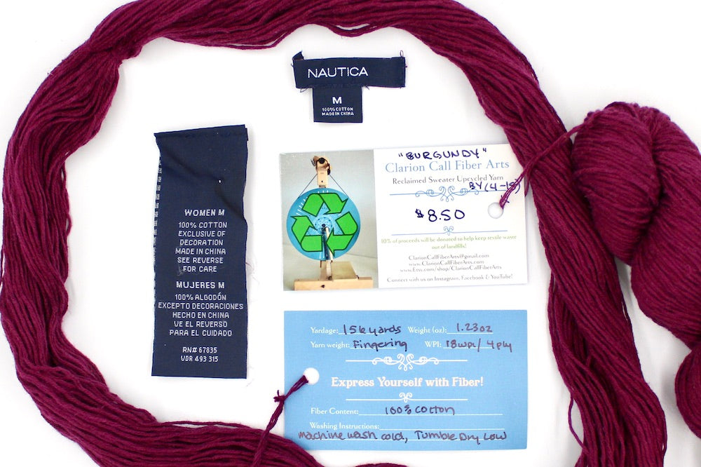A skein of Maroon, Fingering weight yarn encircling the sweater tags it was recycled from, as well as the hand written yarn tags detailing the specs of this upcycled yarn to help you make your next project! 