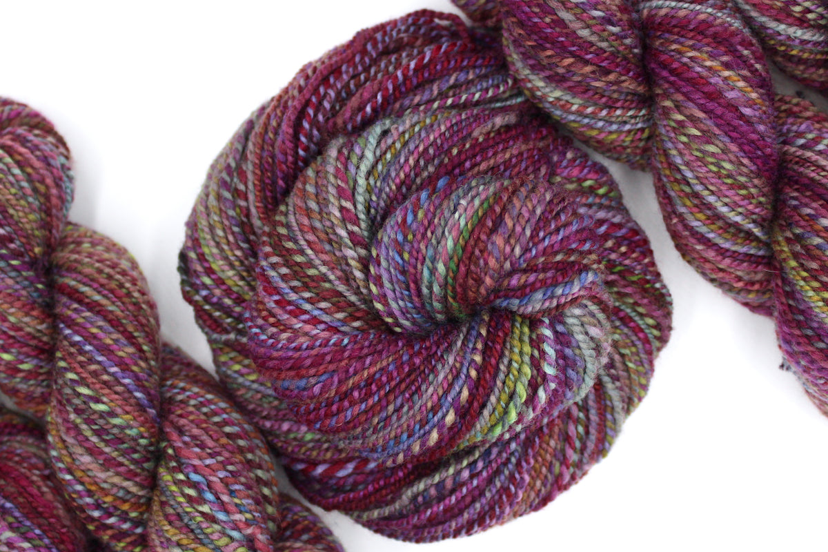 A one of a kind, hand dyed Variegated skein of multicolored Maroon, Pink, Cornflower Blue, and Lime Green self-striping wool Yarn coiled attractively in the center of the frame. 
