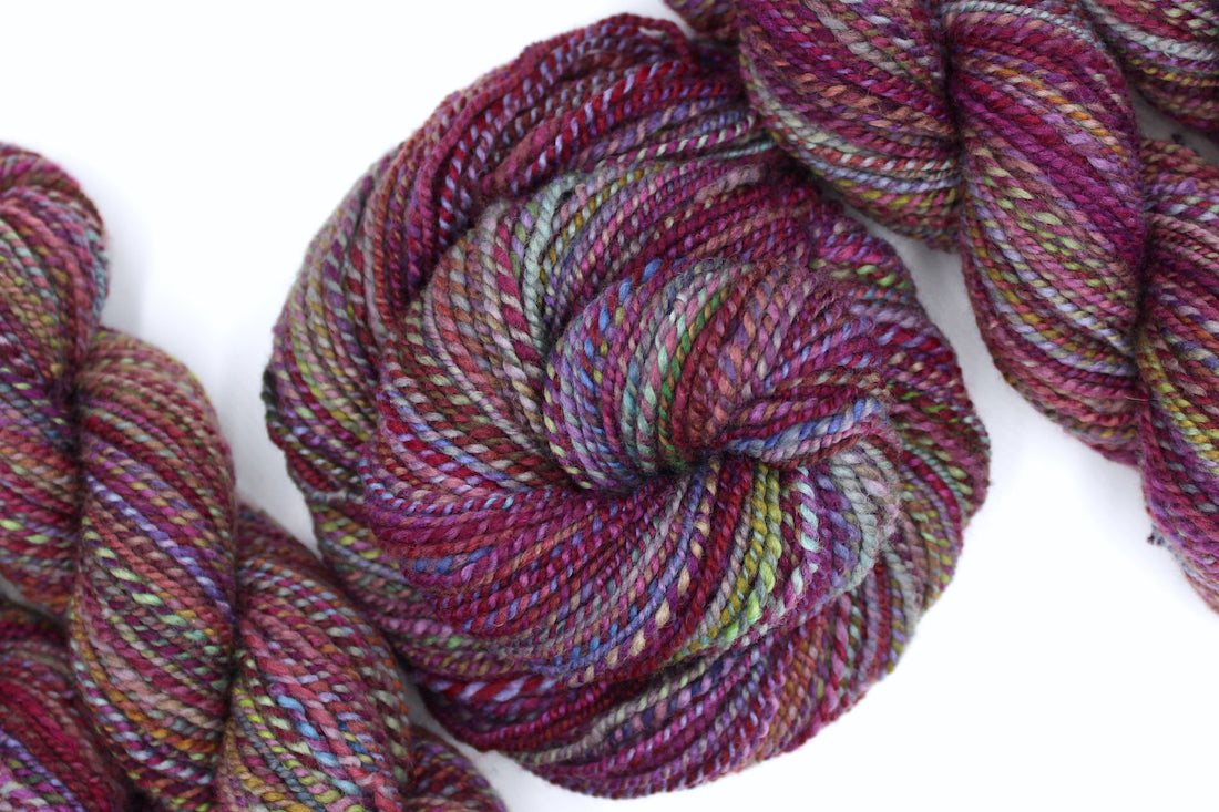 A one of a kind, Dk Weight, hand dyed Variegated skein of self striping Maroon, Pink, Cornflower Blue, and Lime Green Yarn, made of 100% Hand Dyed Merino Wool and coiled attractively in the center of the frame. 