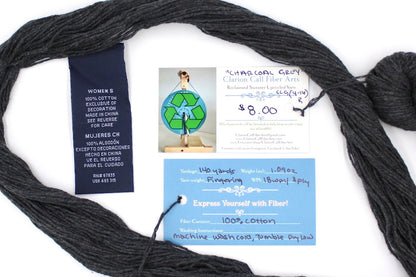 A skein of Dark Grey, Fingering weight yarn encircling the sweater labels it was recycled from, as well as the hand written yarn tags detailing the specs of this upcycled yarn to help you make your next project! 
