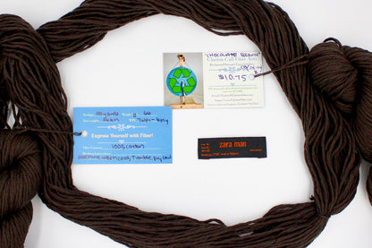 A skein of Vegan, Dark Chocolate Brown, 100% Upcycled Cotton, Heavy Worsted (or Aran) Weight yarn encircling the sweater labels it was recycled from, as well as the hand written yarn tags detailing the specs of this recycled yarn to help you make your next project! 