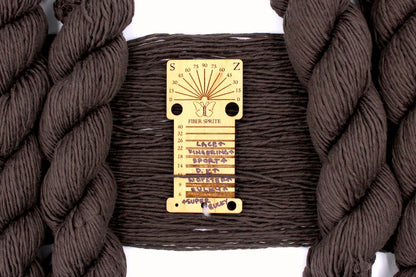 A skein of Vegan, Dark Chocolate Brown, 100% Upcycled Cotton, Heavy Worsted (or Aran) Weight Yarn recycled by hand from unwanted sweaters draped over the center of the frame with one strand on top of a wooden Yarn weight tool, to better illustrate the thickness of the yarn. 