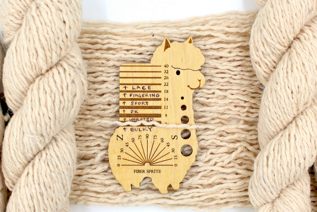 A skein of Beige or Cream, 90% Upcycled Acrylic, 10% Wool, Heavy Worsted (or Aran) Weight Yarn recycled by hand from unwanted sweaters draped over the center of the frame with one strand on top of a Llama shaped Yarn weight tool, to better illustrate the thickness of the yarn. 