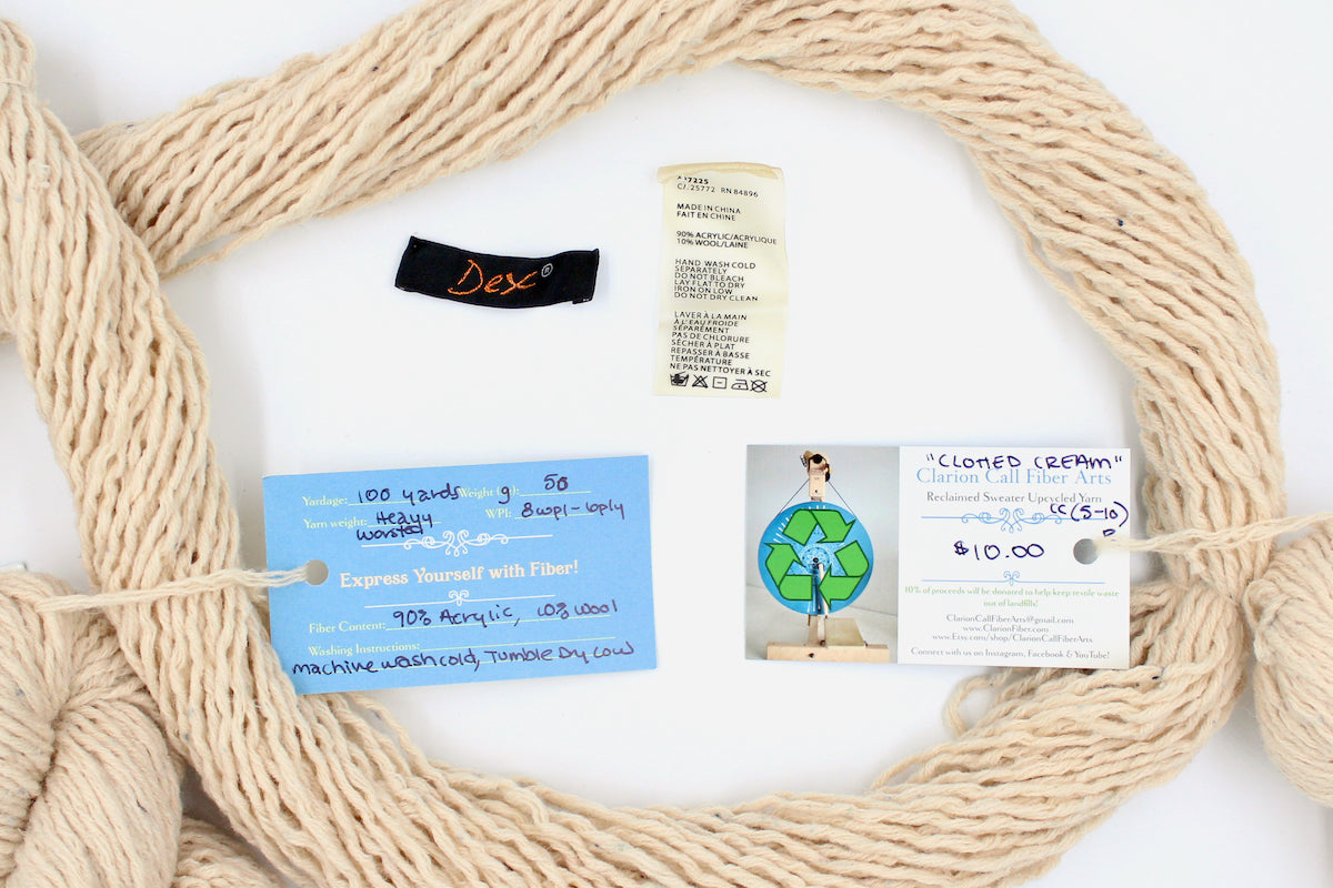A skein of Beige or Cream, 90% Upcycled Acrylic, 10% Wool, Heavy Worsted (or Aran) Weight yarn encircling the sweater labels it was recycled from, as well as the hand written yarn tags detailing the specs of this recycled yarn to help you make your next project! 