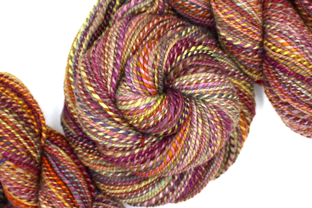 A one of a kind, Sport Weight, hand dyed variegated skein of multicolored Red, Orange, Yellow, and Purple self-striping 100% Merino wool Yarn coiled attractively in the center of the frame. 