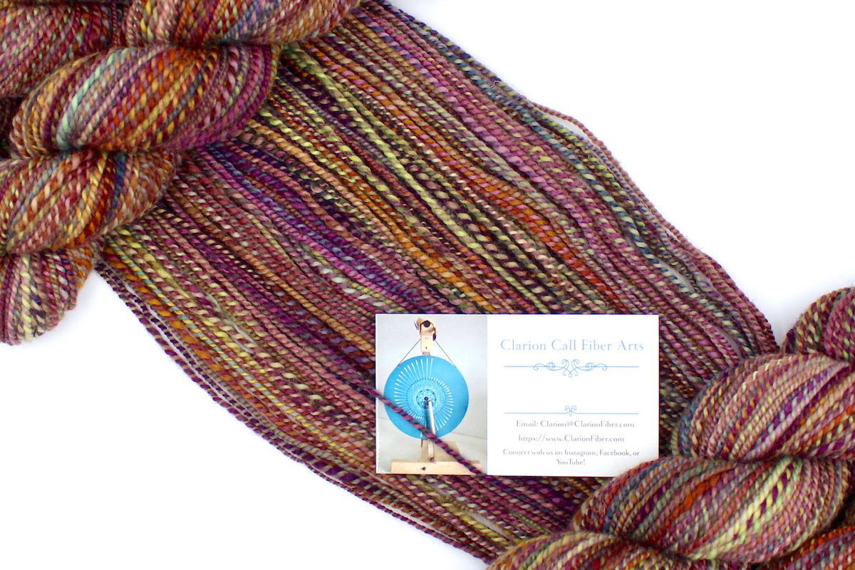 A one of a kind, Sport Weight, hand dyed variegated skein of multicolored Red, Orange, Yellow, and Purple self-striping 100% Merino wool Yarn draped diagonally across the frame, so you can really see the color play. 