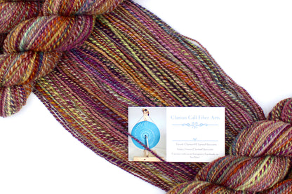 A one of a kind, Sport Weight, hand dyed variegated skein of multicolored Red, Orange, Yellow, and Purple self-striping 100% Merino wool Yarn draped diagonally across the frame, so you can really see the color play. 