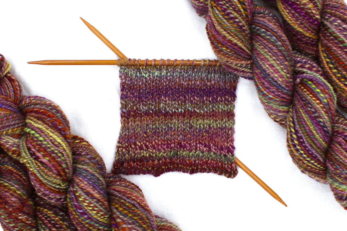 A sample swatch knitted from a one of a kind, Sport Weight, hand dyed variegated skein of multicolored Red, Orange, Yellow, and Purple self-striping 100% Merino wool Yarn. 