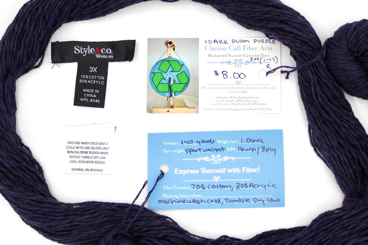 A skein of Dark Purple, Sport yarn encircling the sweater labels it was recycled from, as well as the hand written yarn tags detailing the specs of this upcycled yarn to help you make your next project! 