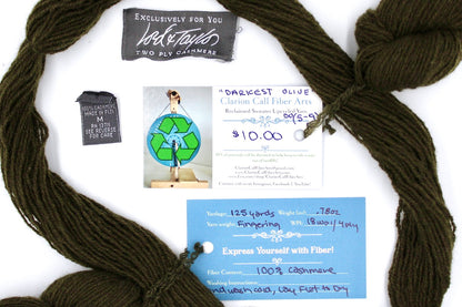A skein of Dark Olive Cashmere, Fingering yarn encircling the sweater labels it was recycled from, as well as the hand written yarn tags detailing the specs of this upcycled yarn to help you make your next project! 