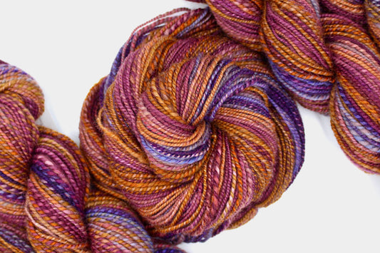 A one of a kind, hand dyed gradient skein of multicolored Light Rust, Mauve Pink, Orange, Gold, Brown, Lavender, and Deep Purple self-striping wool Yarn coiled attractively in the center of the frame. 