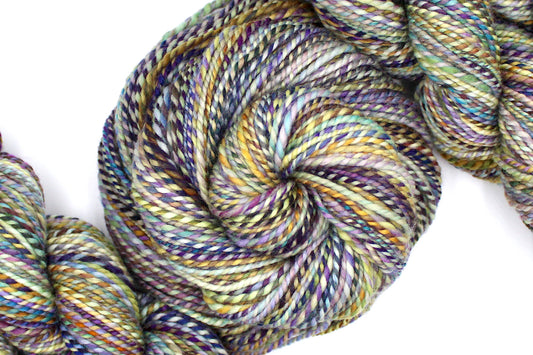 A one of a kind, Dk Weight, hand dyed variegated skein of multicolored Navy, Gold, and Purple self-striping 100% Superwash Merino wool Yarn coiled attractively in the center of the frame. 