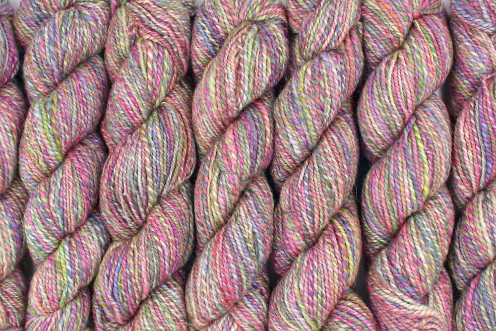 Six one of a kind, Dk Weight, hand dyed variegated skeins of multicolored Pink, Green, Yellow, Blue, and Purple self-striping Hand spun Yarn, made of Hand Dyed Wool and Sparkly Nylon Fiber, lined up side by side. 