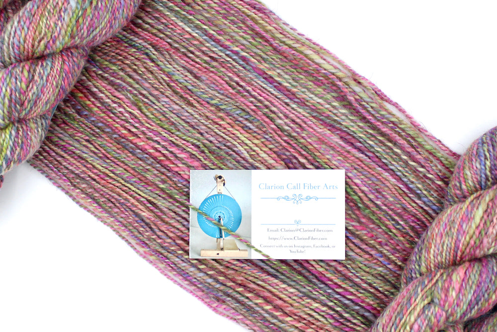 A one of a kind, Dk Weight, hand dyed variegated skein of multicolored Pink, Green, Yellow, Blue, and Purple self-striping Hand spun Yarn, made of Hand Dyed Wool and Sparkly Nylon Fiber, draped diagonally across the frame, so you can really see the color play. 