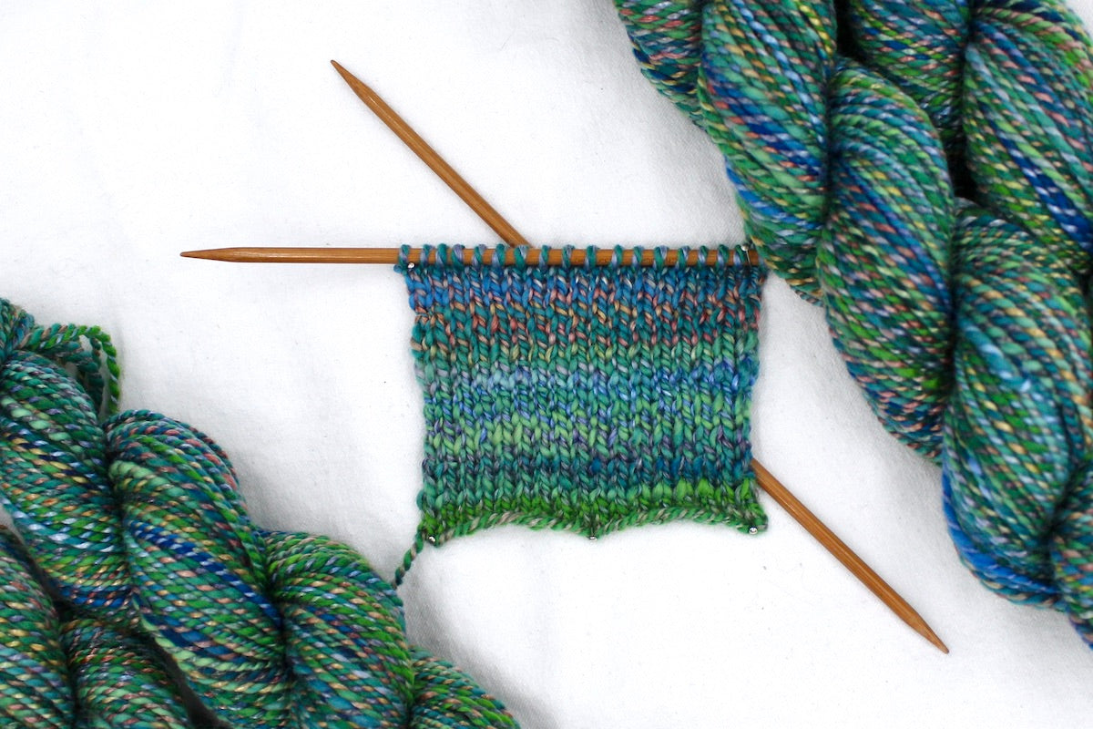 A sample swatch knitted from a one of a kind, hand dyed variegated skein of multicolored Green, Blue, Gold, and Red self-striping wool Yarn. 