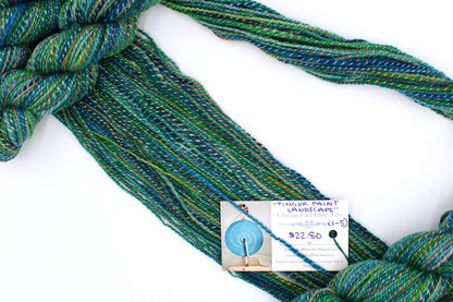 A one of a kind, hand dyed gradient skein of variegated skein of multicolored Green, Blue, Gold, and Red self-striping wool Yarn draped diagonally across the frame, so you can really see the color play. 