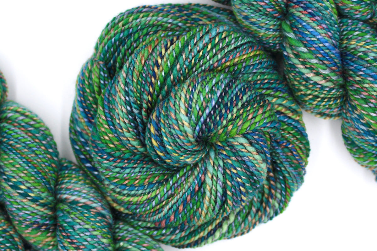 A one of a kind, hand dyed variegated skein of multicolored Green, Blue, Gold, and Red self-striping wool Yarn coiled attractively in the center of the frame. 