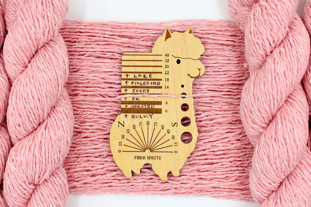 A skein of Vegan, Pink, 60% Cotton, 40% Acrylic, Dk Weight Yarn recycled by hand from unwanted sweaters draped over the center of the frame with one strand on top of a Llama shaped Yarn weight tool, to better illustrate the thickness of the yarn. 