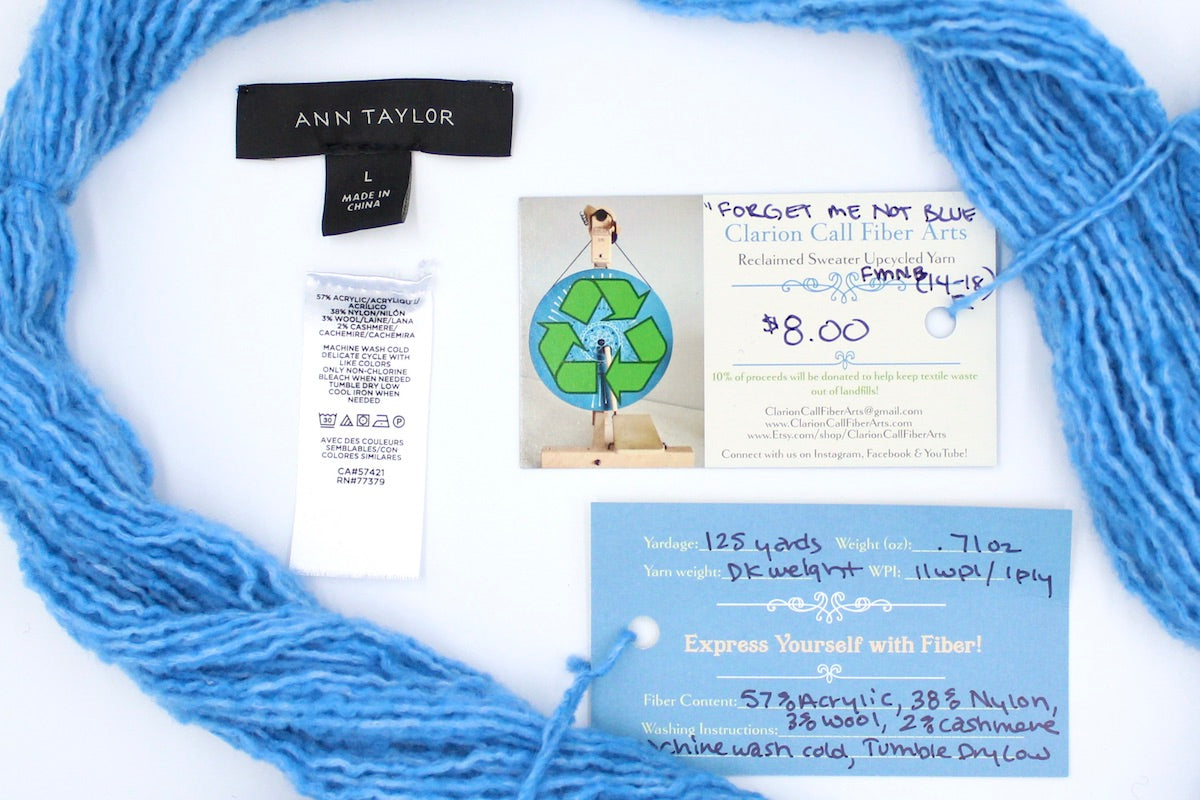 A skein of Light Baby Blue, Acrylic, Nylon, Wool, Cashmere, Dk Weight yarn encircling the sweater labels it was recycled from, as well as the hand written yarn tags detailing the specs of this upcycled yarn to help you make your next project! 