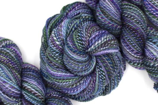 A one of a kind, DK Weight, hand dyed variegated skein of multicolored Purple, Lavender, Forest Green, Baby Blue, and Light Teal, self-striping wool Yarn coiled attractively in the center of the frame.