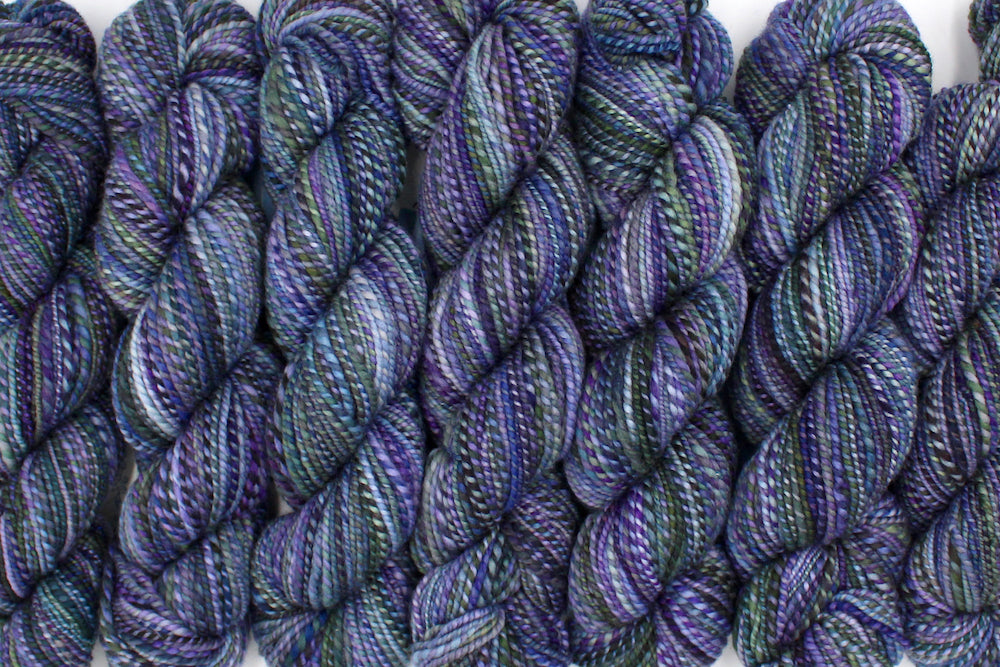 Six one of a kind, Dk weight, hand dyed variegated skein of multicolored Purple, Lavender, Forest Green, Baby Blue, and Light Teal self-striping wool Yarn lined up side by side. 