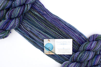 A one of a kind, Dk weight, hand dyed variegated skein of multicolored Purple, Lavender, Forest Green, Baby Blue, and Light Teal self-striping wool Yarn draped diagonally across the frame, so you can really see the color play. 