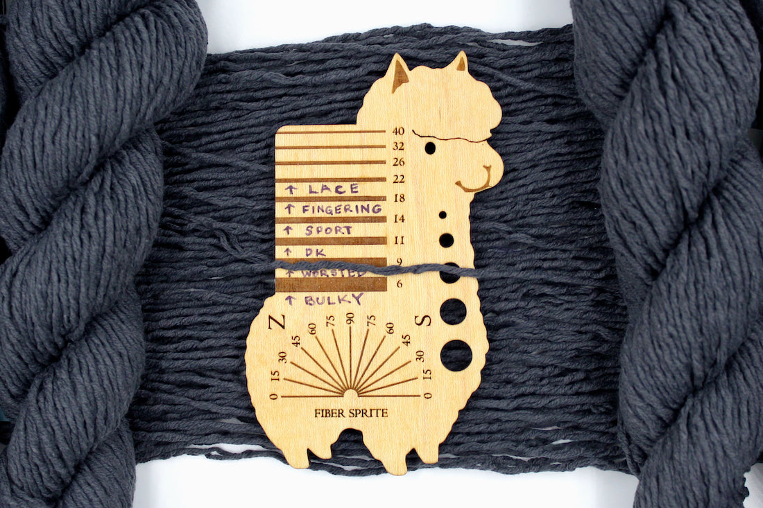 A skein of Vegan, Dark Grey, 60% Cotton, 40% Acrylic, Worsted Weight Yarn recycled by hand from unwanted sweaters draped over the center of the frame with one strand on top of a Llama shaped Yarn weight tool, to better illustrate the thickness of the yarn. 