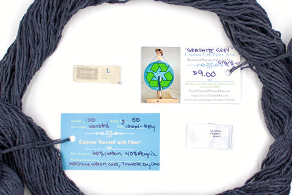 A skein of Vegan, Dark Grey, 60% Cotton, 40% Acrylic, Worsted Weight yarn encircling the sweater labels it was recycled from, as well as the hand written yarn tags detailing the specs of this recycled yarn to help you make your next project! 
