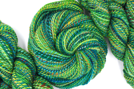 A one of a kind, hand dyed variegated skein of multicolored Green, Blue and Yellow self-striping wool Yarn coiled attractively in the center of the frame. 