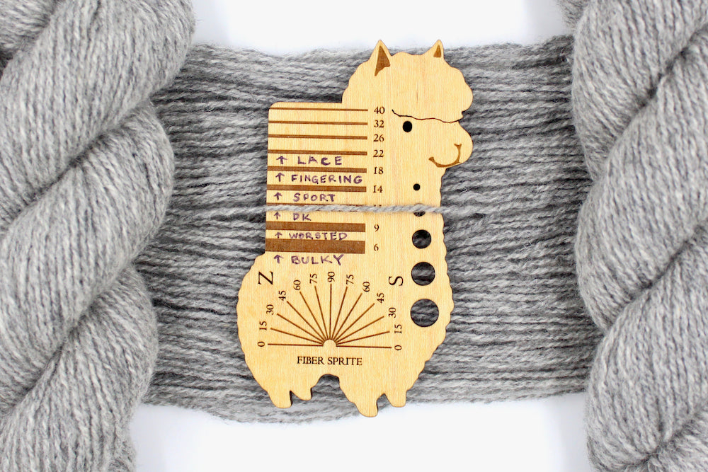 A skein of Light heathered grey, 90% Lambs wool and 10% cashmere, Dk Weight Yarn recycled by hand from unwanted sweaters draped over the center of the frame with one strand on top of a Llama shaped Yarn weight tool, to better illustrate the thickness of the yarn. 